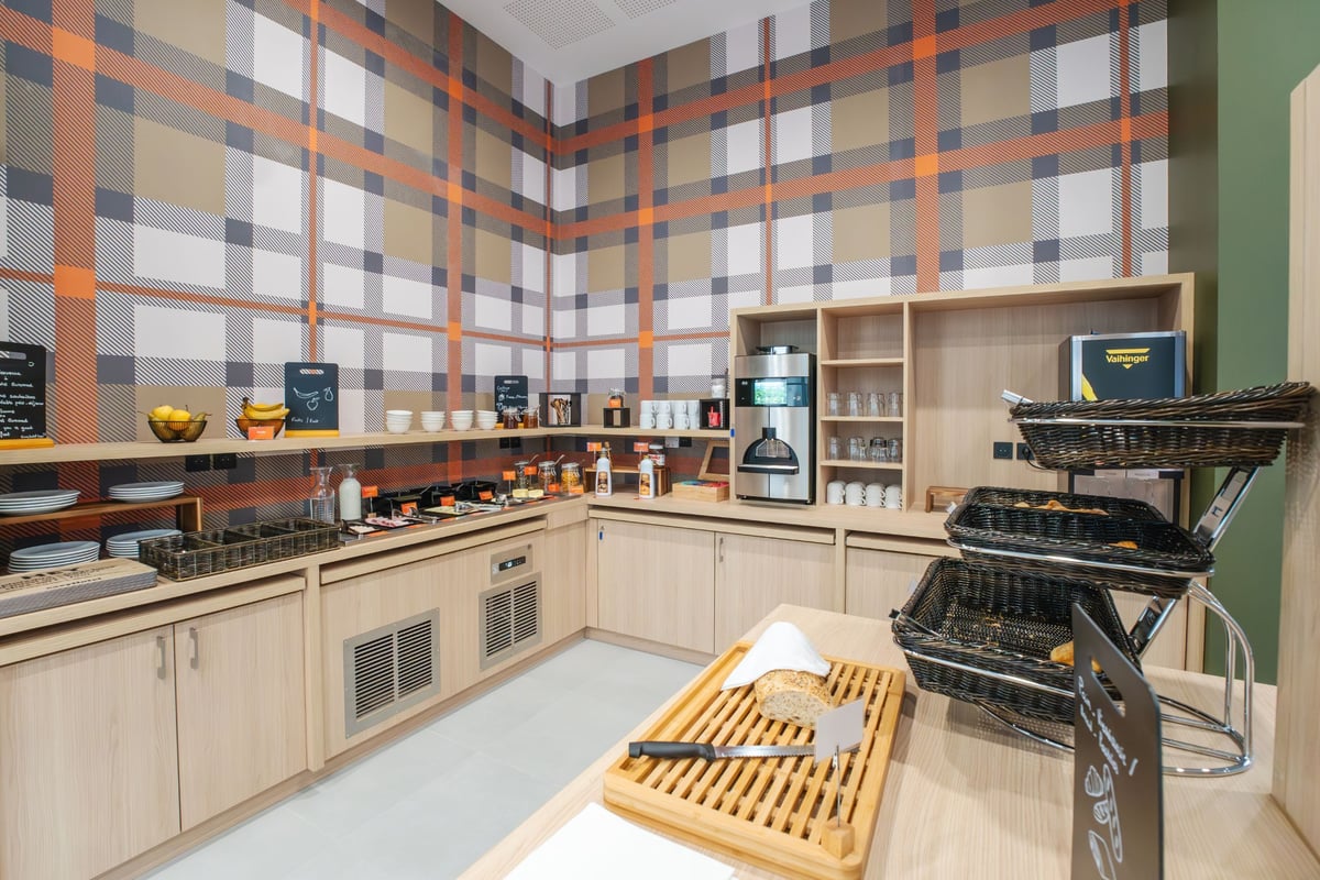 easyHotel Marseille Euromed breakfast room featuring a modern buffet with fresh bread, fruit, pastries, and coffee, against a vibrant orange and gray plaid wall.