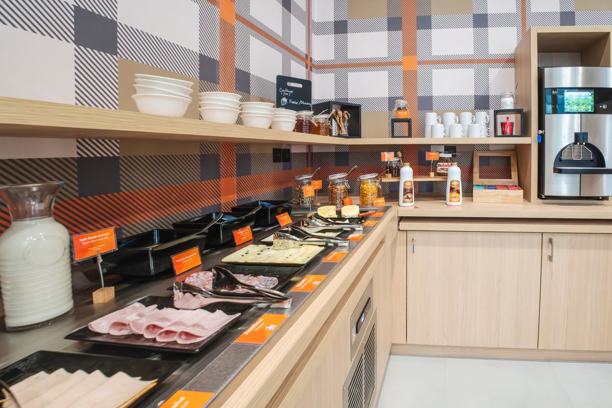 easyHotel Marseille Euromed breakfast room with a full buffet, including cold meats, cheeses, milk, cereals, and a coffee machine, set against an orange, grey, and white plaid wall.