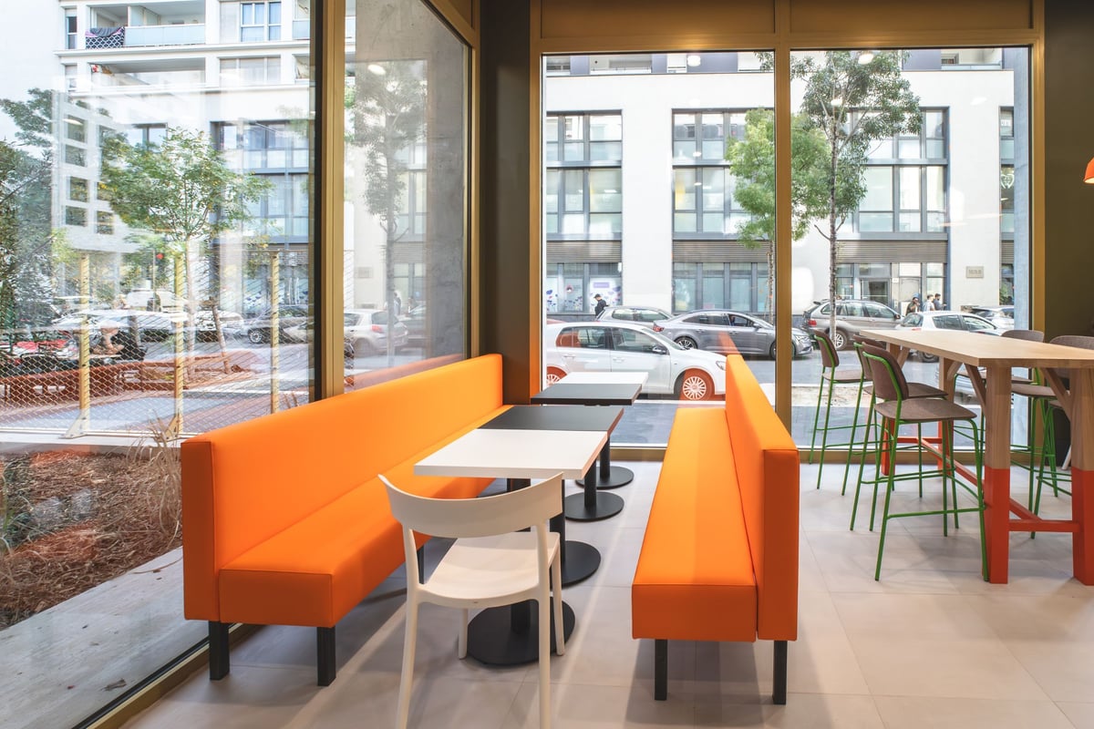 easyHotel Marseille Euromed breakfast room with vibrant orange booth seating, a tall wooden communal table with green-framed bar stools, and large windows overlooking an urban street.