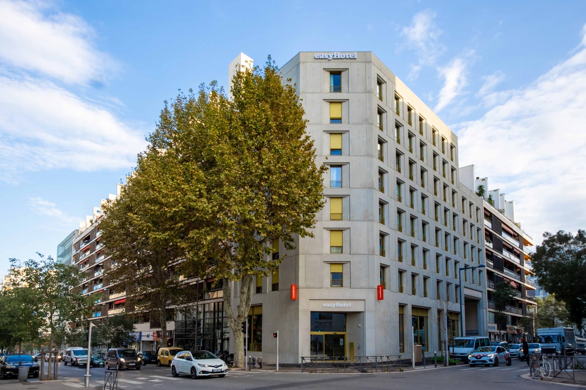 Exterior view of the easyHotel Marseille Euromed, a modern multi-story hotel building with a light-colored facade and yellow window accents, situated on a city street corner with trees and vehicles.