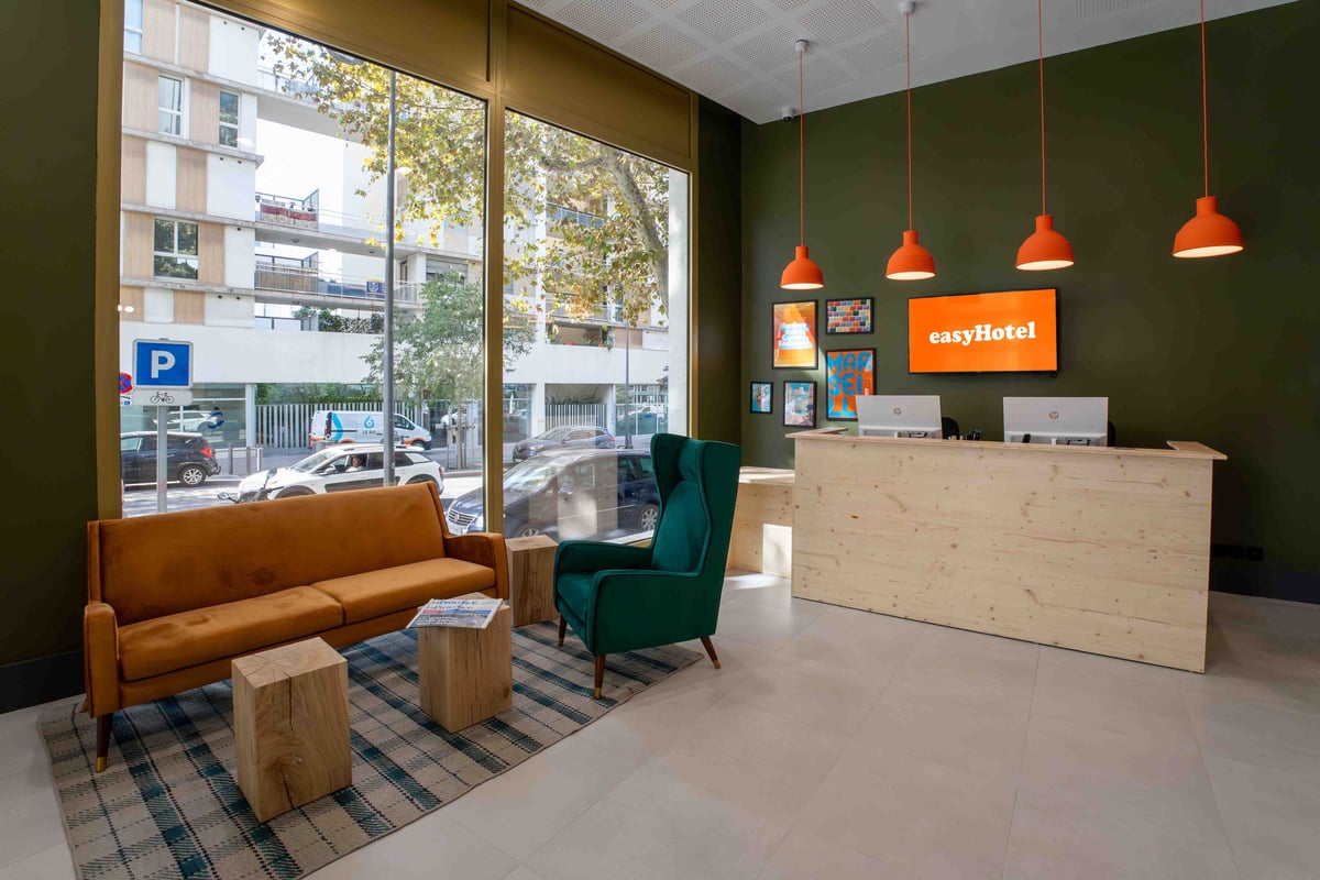 Modern easyHotel Marseille Euromed reception area featuring a light wood desk, dark green walls, bright orange pendant lights, an orange sofa, and a green armchair, with a city street visible through large windows.