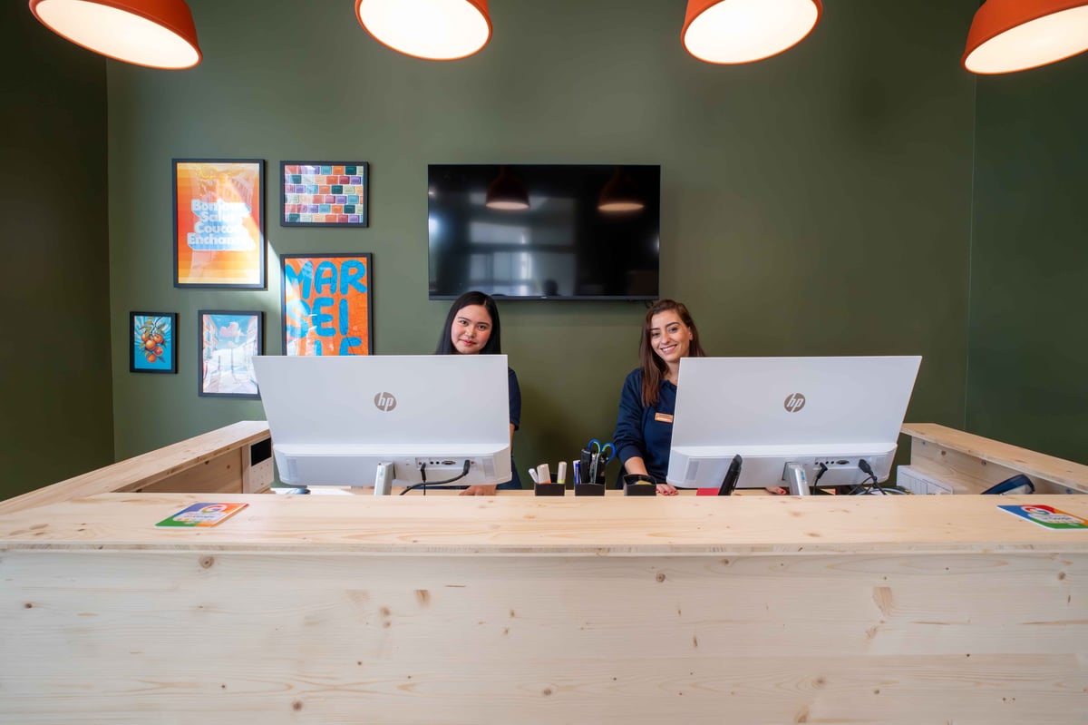 easyHotel Marseille Euromed reception desk with two smiling staff members behind a light wood counter, set against a dark green wall decorated with local art.