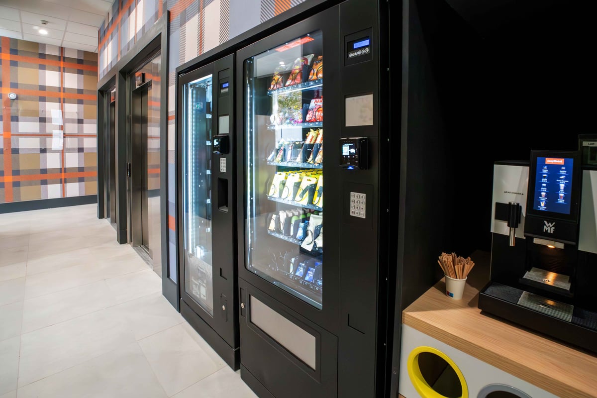 EasyHotel Marseille Euromed lobby featuring two black vending machines with snacks and drinks, a self-service coffee machine on a wooden counter, and a distinctive plaid wall pattern.