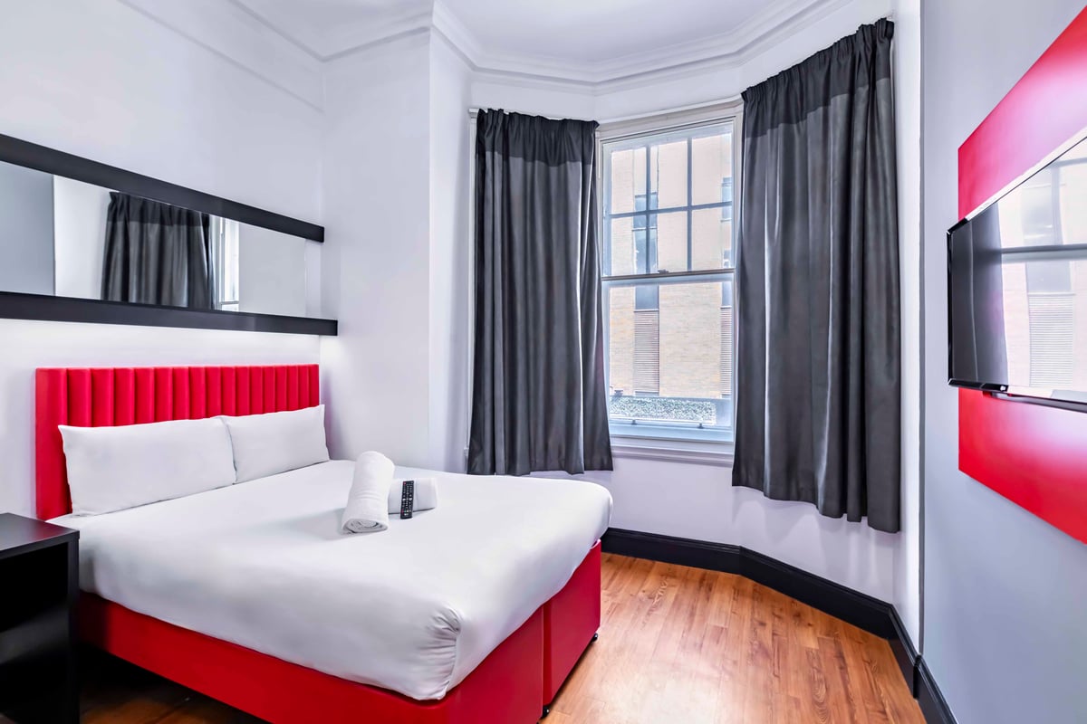 EasyHotel Newcastle Superior Double room with a double bed, white linens, red headboard and bed frame, and a window with grey curtains.