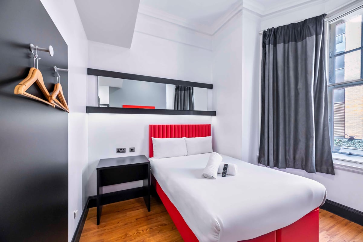EasyHotel Newcastle Superior Double room with double bed, white linens, red headboard, bedside table, and window with grey curtains.