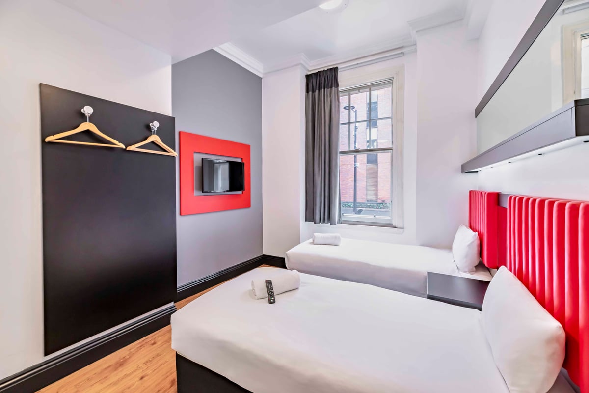 EasyHotel Newcastle Twin Room with two single beds, white linens, red headboards, and a window with gray curtains.