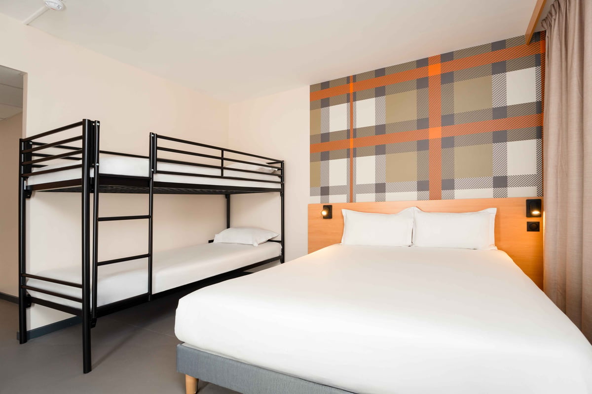 easyHotel Nice Old Town family room featuring a double bed and black metal bunk beds with white linens. The room has a modern plaid accent wall in orange, grey, and white, and a wooden headboard with integrated lighting.