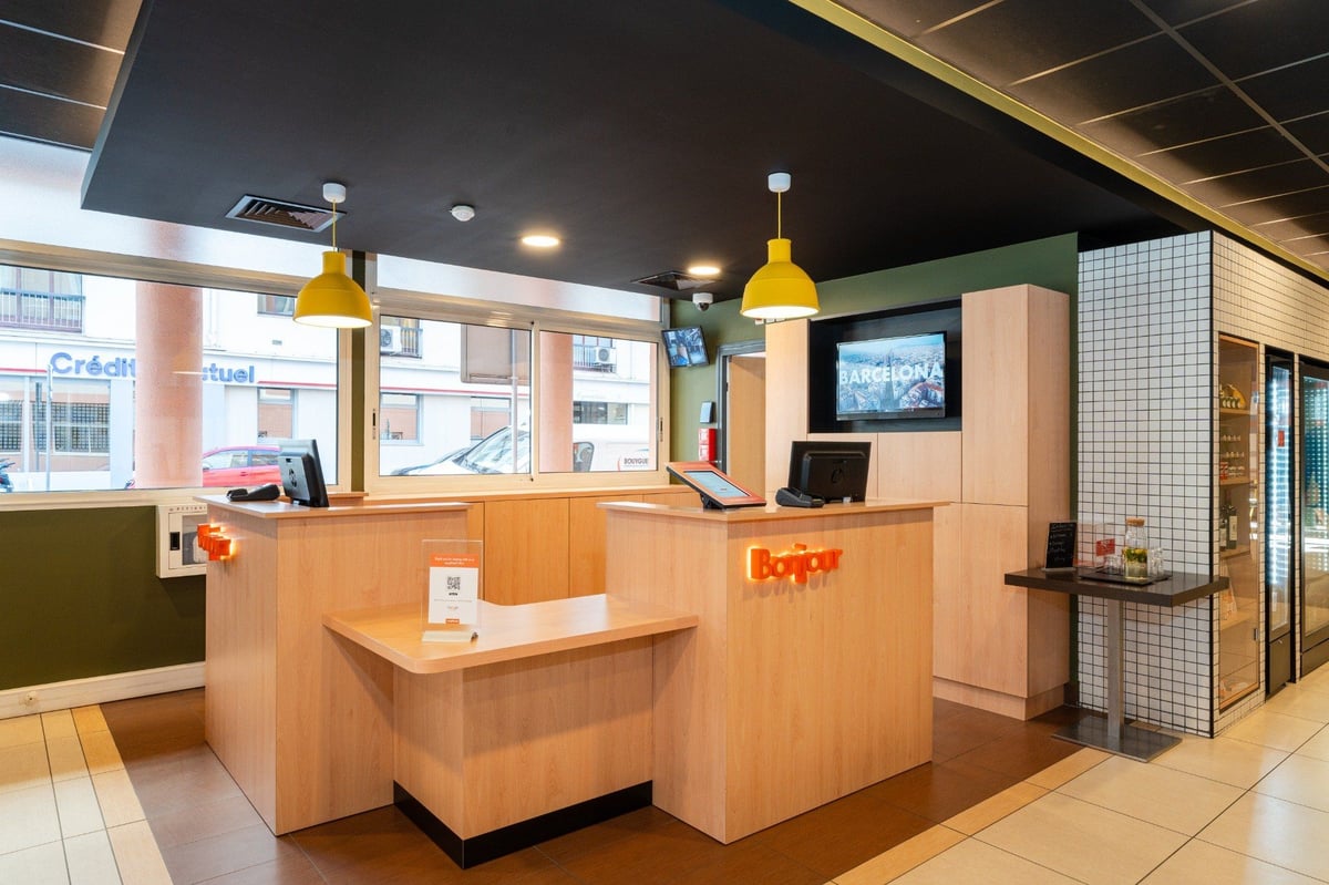 EasyHotel Nice Old Town reception area featuring a modern wooden front desk with an orange 'Bonjour' sign, yellow pendant lights, a large screen, and a tiled convenience store section.