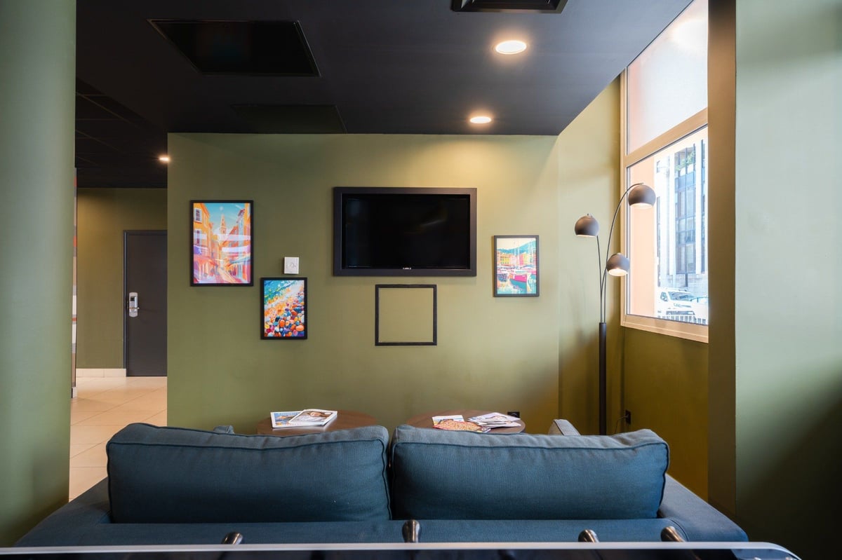 easyHotel Nice Old Town reception lounge with olive green walls, a dark blue sofa, wall-mounted TV, vibrant framed artwork, and a large window.
