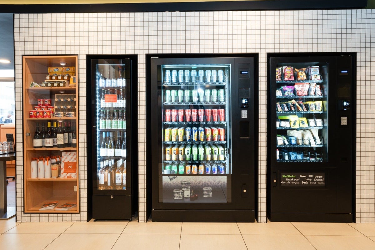 ALT: A modern self-service mini-market at easyHotel Nice Old Town, featuring a wooden display case with wine and snacks, and multiple black vending machines offering bottled drinks, colorful canned sodas, and various packaged snacks, set against a white g