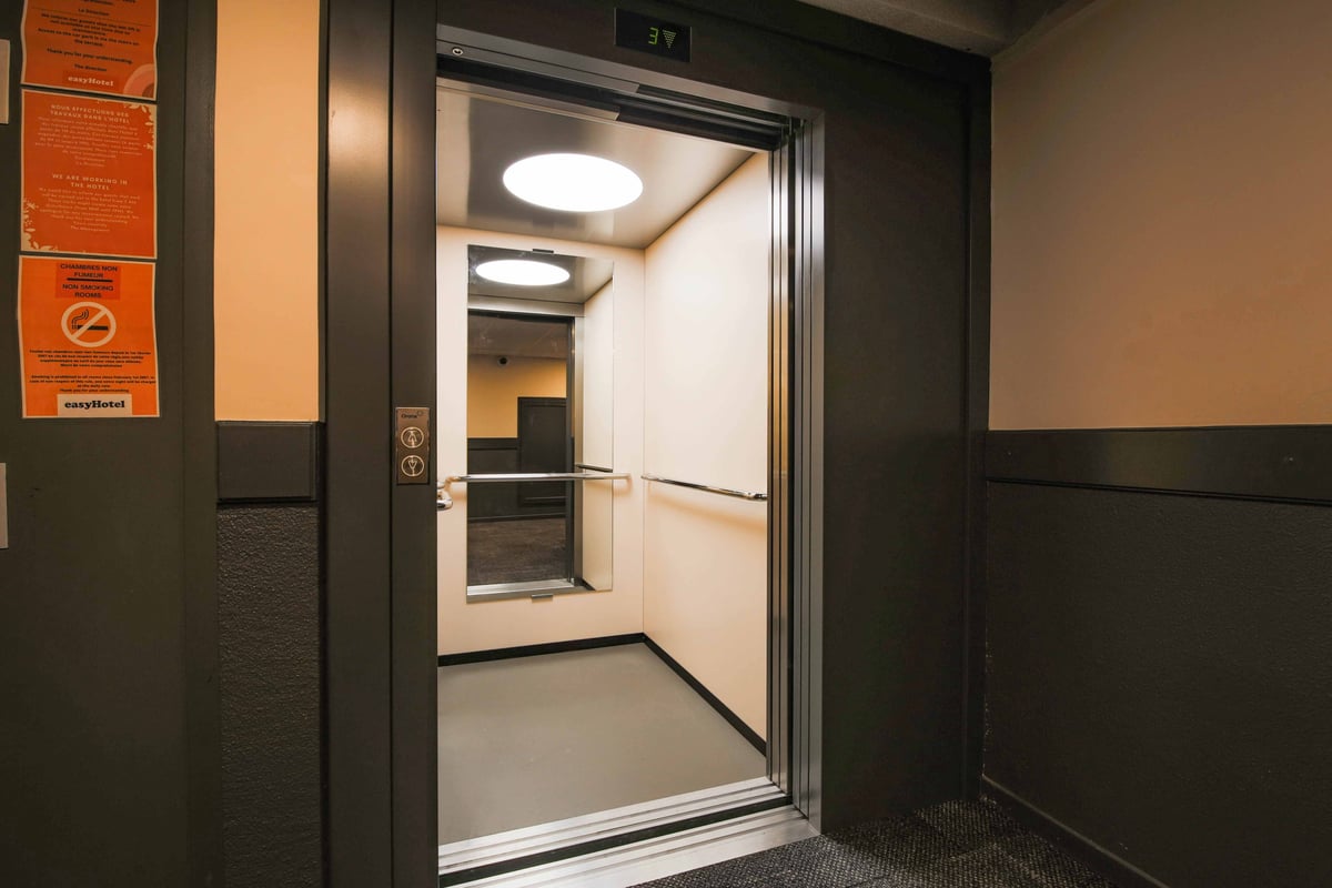 An open, modern elevator with reflective surfaces and handrails, located in the dark hallway of an easyHotel Nice. EasyHotel branding and no-smoking signs are visible on the adjacent wall.