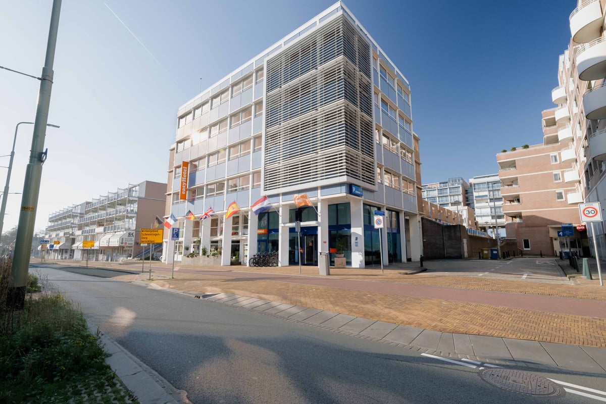 Exterior view of the easyHotel Scheveningen Beach, a modern multi-story building with a light blue and white facade, situated on a street under a clear sky.