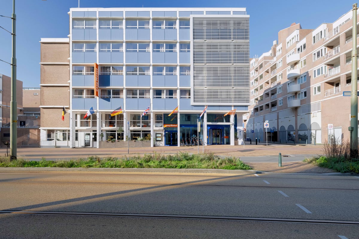 Exterior view of the easyHotel Scheveningen Beach, a modern light blue building with white window frames, under a clear sky. European flags are displayed in front of the entrance, with a street and tram tracks in the foreground.
