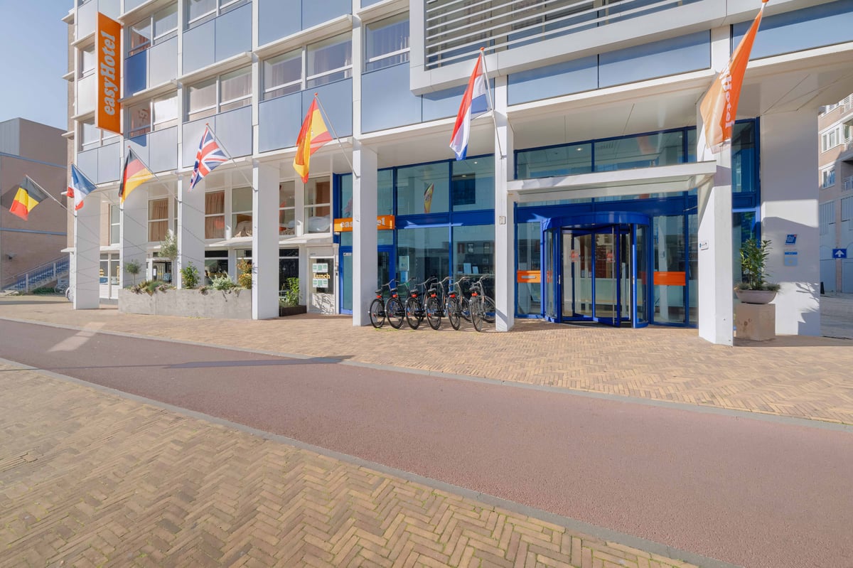 The modern exterior of easyHotel Scheveningen Beach, featuring its prominent orange banner, blue glass entrance with a revolving door, and international flags. Several bicycles are parked near the entrance.