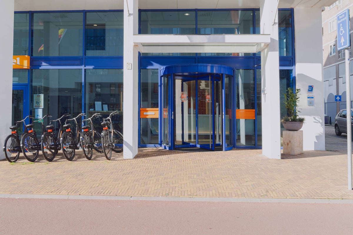Exterior view of easyHotel Scheveningen Beach, featuring its distinctive blue-framed entrance with a revolving door, white columns, and multiple bicycles parked in front.
