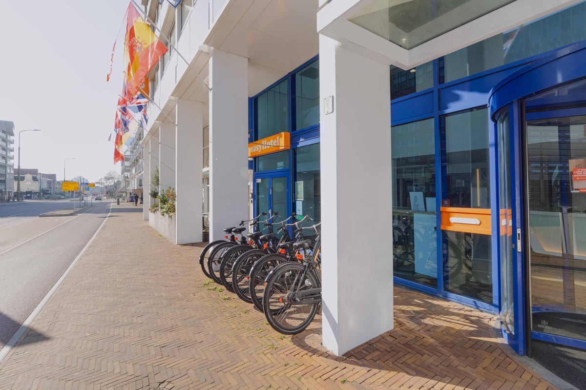 easyHotel Scheveningen Beach exterior featuring its orange sign, blue entrance, white pillars, parked bicycles, and international flags, alongside a brick sidewalk.