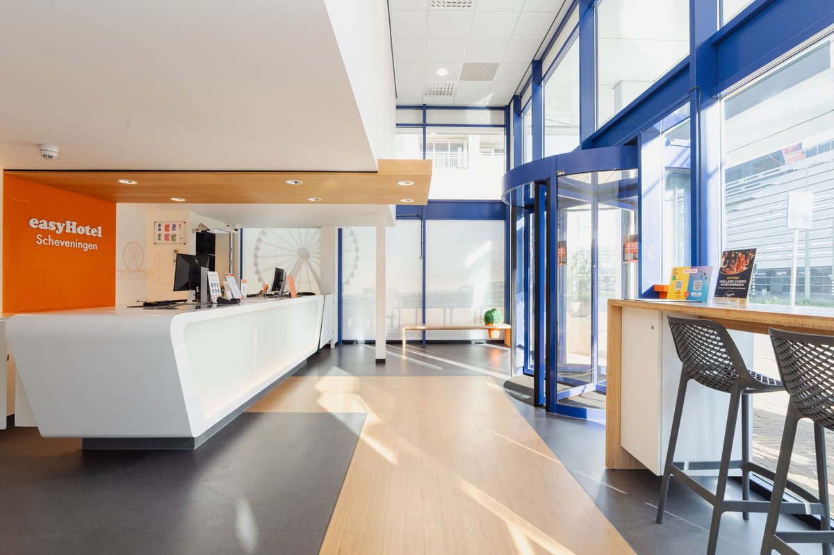 Bright, modern reception lobby at easyHotel Scheveningen Beach, featuring a long white desk, an orange branded wall, and large windows with a revolving door and a ferris wheel graphic.