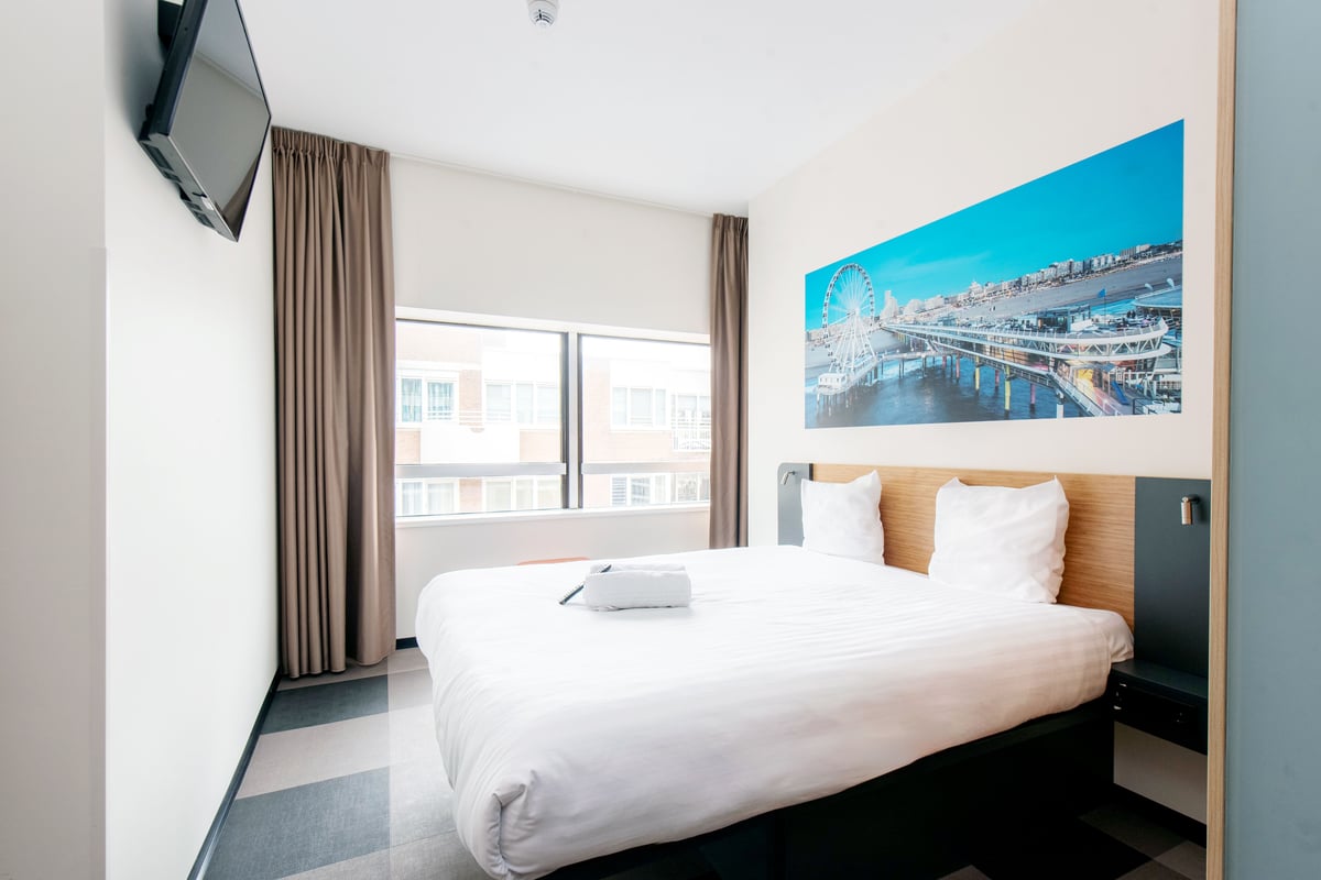 An easyHotel Scheveningen Beach Superior Double room featuring a comfortable double bed, a wall-mounted TV, and a large vibrant mural of the iconic Scheveningen Pier and Ferris Wheel.