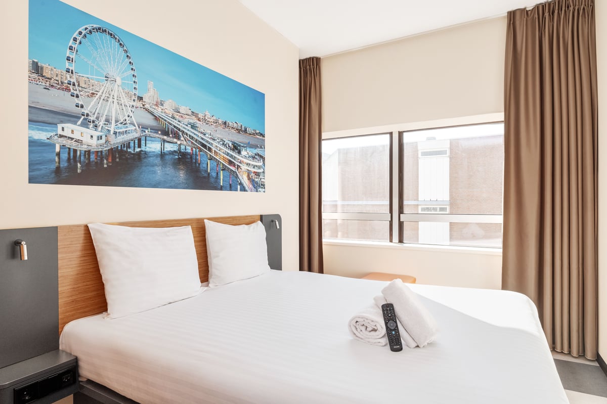 A bright, modern easyHotel room features a neatly made double bed with white linens, two white folded towels, and a TV remote. A large mural above the bed displays the iconic Ferris wheel on Scheveningen Pier with the beach and cityscape in the background
