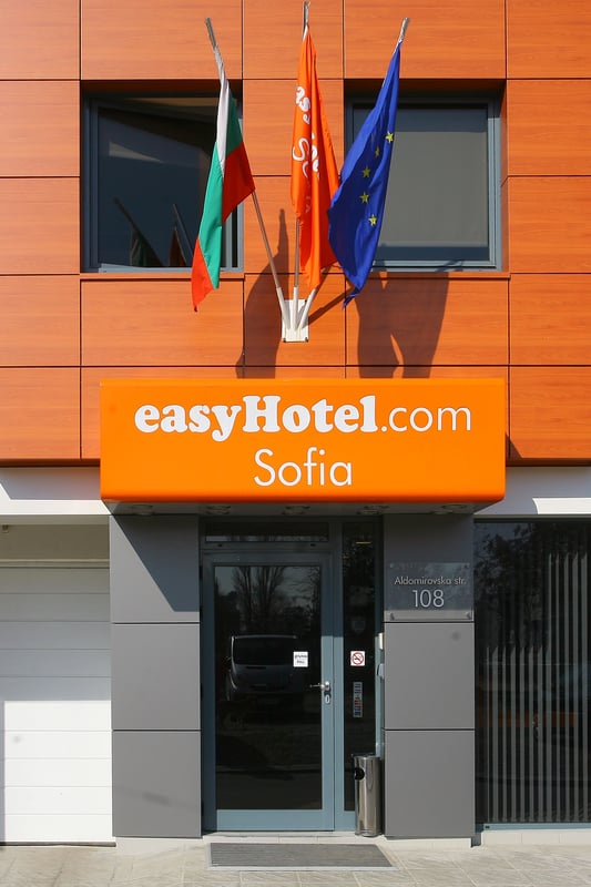 Exterior view of easyHotel Sofia, showcasing the bright orange easyHotel.com sign, the Bulgarian flag, and the European Union flag on its modern facade.