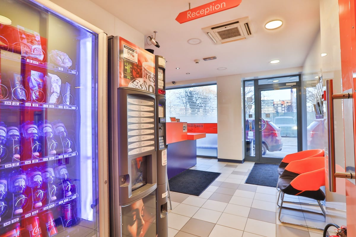 Modern easyHotel Sofia reception area with vibrant orange design elements. The lobby features self-service vending machines for drinks, snacks, and coffee, a contemporary reception desk, and a bright waiting area with orange chairs near the glass entrance
