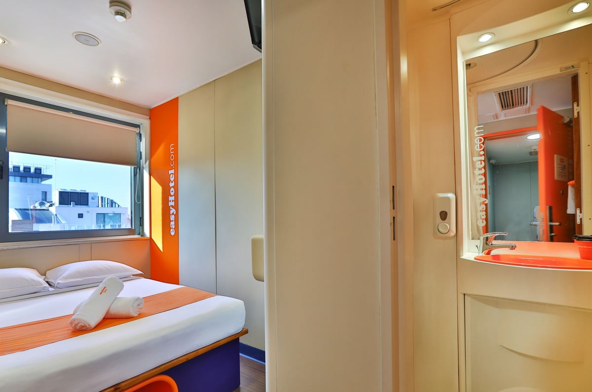 easyHotel Sofia small double room with a compact bed, orange accents, a large window, and an integrated en-suite bathroom with a sink.