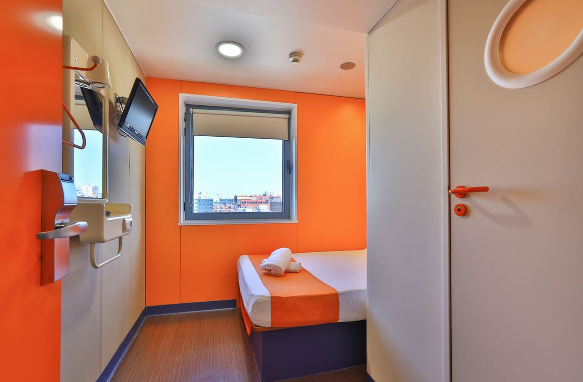 EasyHotel Sofia small double room featuring vibrant orange walls, a compact bed with white and orange bedding, two rolled towels, a wall-mounted television, a minimalist sink area, and a window offering a city view.