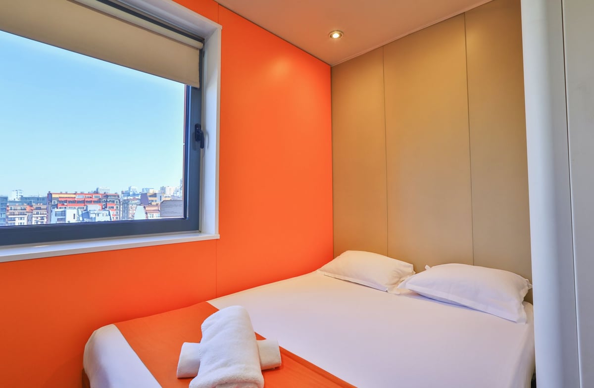 easyHotel Sofia small double room with a minimalist design, featuring a white small double bed on an orange base against a bright orange wall, and a window with a city view of Sofia. White rolled towels are placed on the bed.