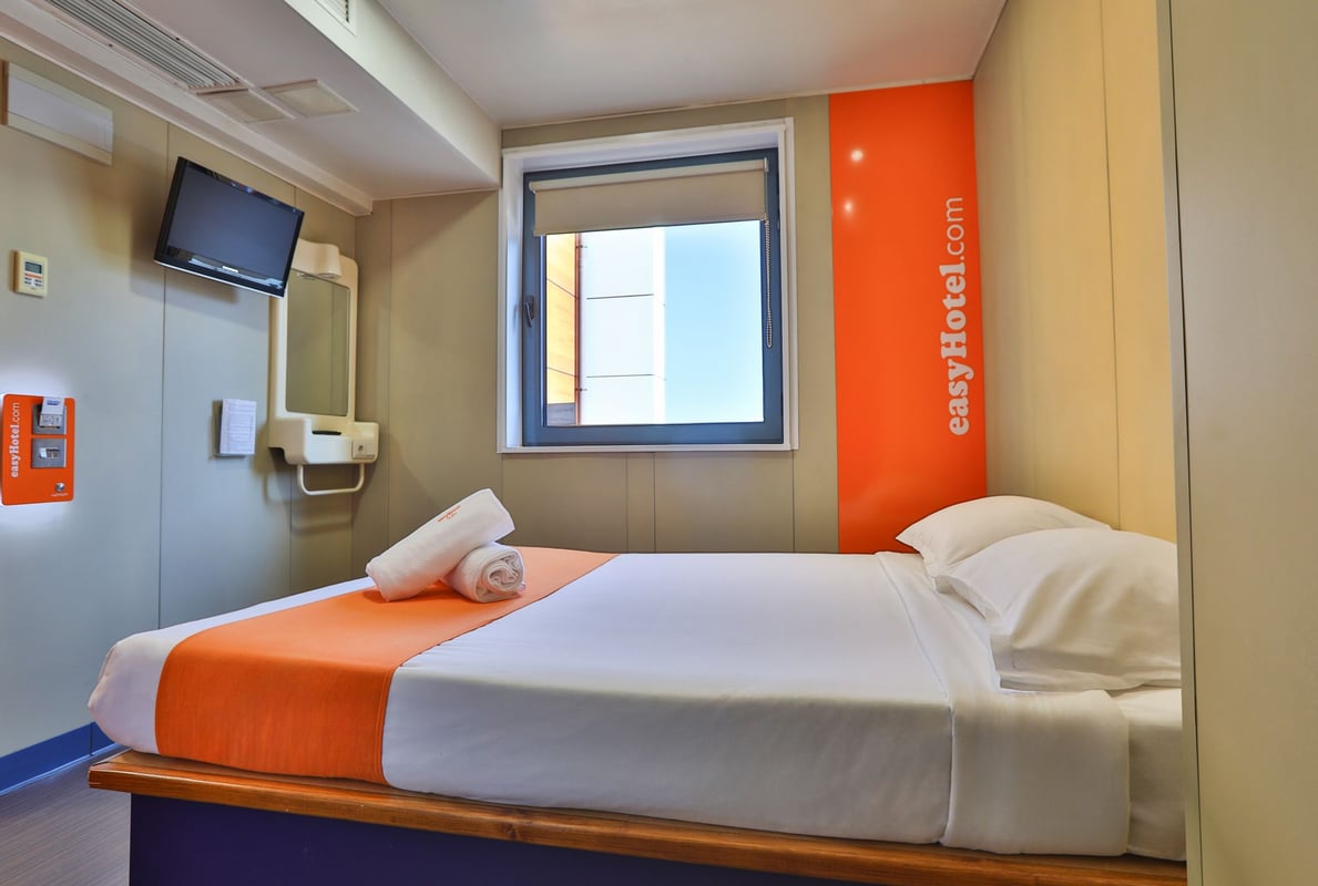 easyHotel Sofia Standard Double room featuring a bed with white bedding, an orange runner, two rolled towels, a window, a wall-mounted TV, and prominent easyHotel branding on an orange wall panel.