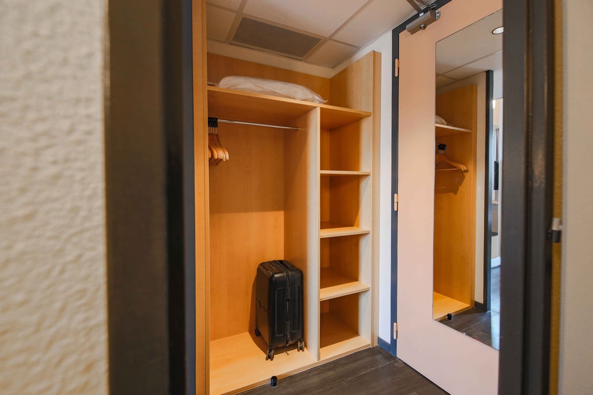An easyHotel room's open wardrobe with light wood shelves, a hanging rail with wooden hangers, and a black suitcase; a mirrored door is visible to the right.