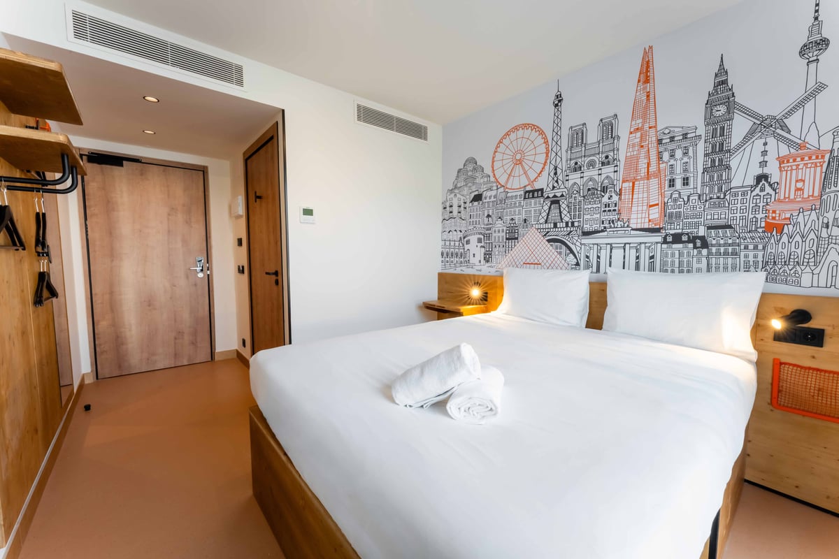 EasyHotel Valencia Ciutat Vella double room with a double bed, white linens, bedside table, and a city skyline mural.