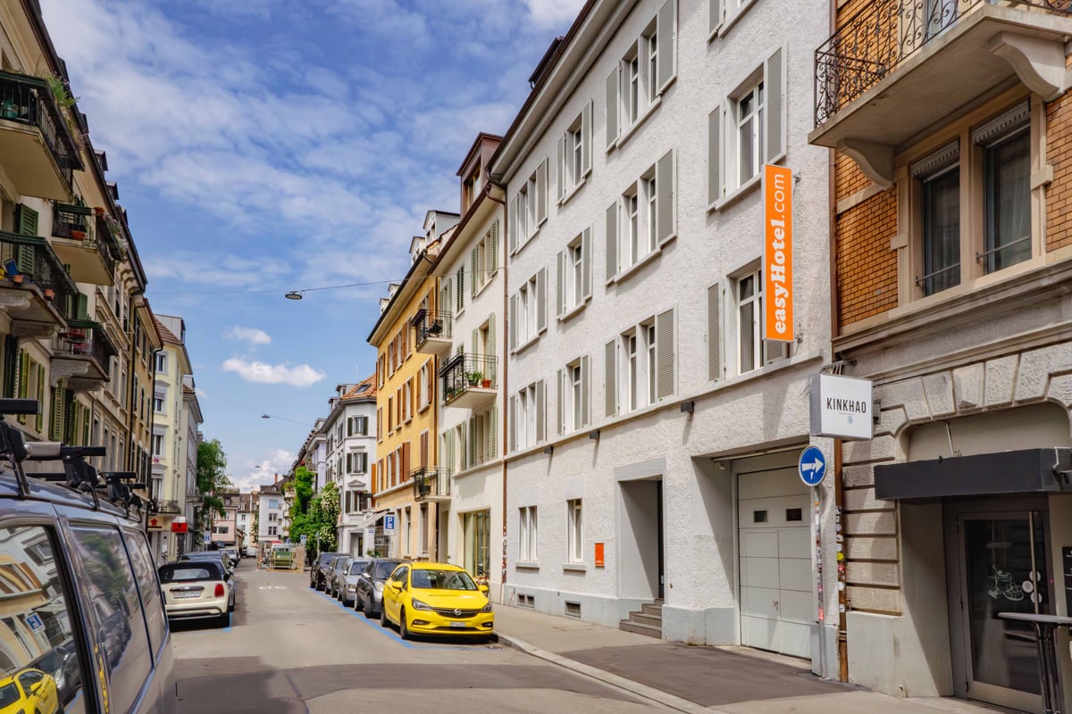 easyHotel Zurich City Centre exterior, a street view of a white building with easyHotel signage, parked cars, and a blue sky.