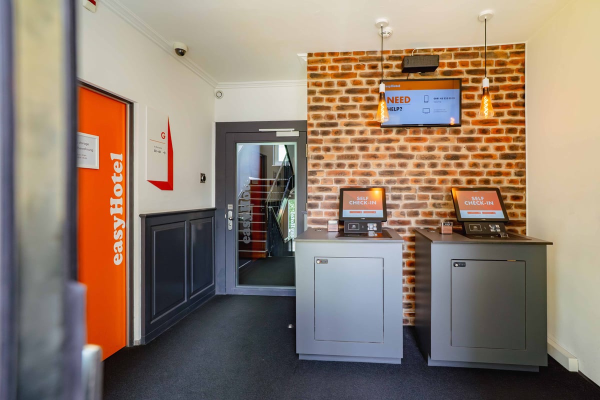 easyHotel Zurich City Centre reception area featuring self-check-in kiosks, a brick wall, and a digital information screen.