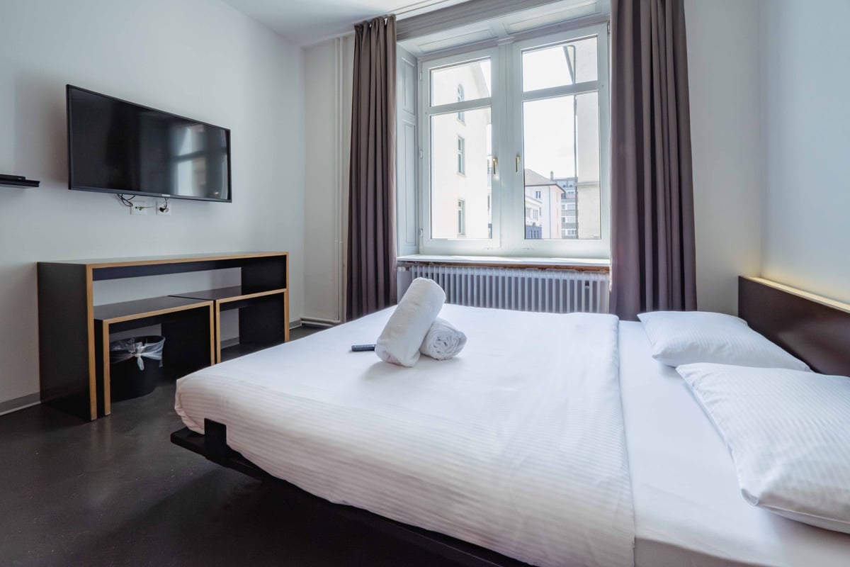 EasyHotel Zurich City Centre Superior Double room with a double bed, white linens, two pillows, a bedside table, a TV mounted on the wall, and a window with brown curtains.