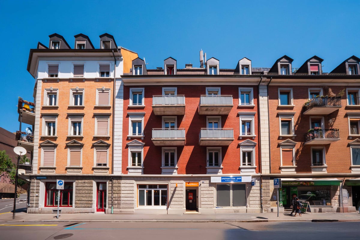 EasyHotel Zurich Limmatplatz exterior, showing a multi-story building with brick and stucco facade, balconies, and street-level shops under a clear blue sky.