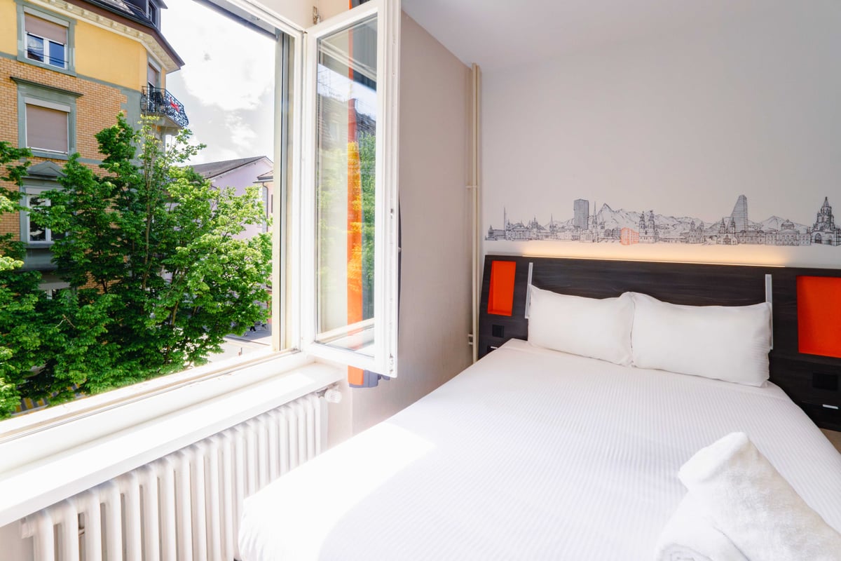 EasyHotel Zurich Main Station double room with white bedding, pillows, and orange headboard accents, featuring a window overlooking a city street and greenery.