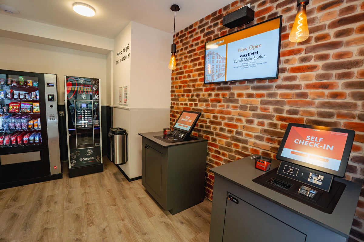 easyHotel Zurich Main Station reception area with self check-in kiosks, vending machines, and a brick accent wall.
