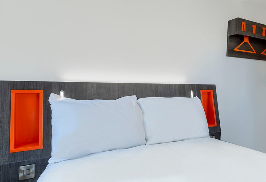 easyHotel Reading room with a bed featuring white linen, a dark grey headboard with orange storage niches, reading lights, and a wall-mounted orange hanger unit.