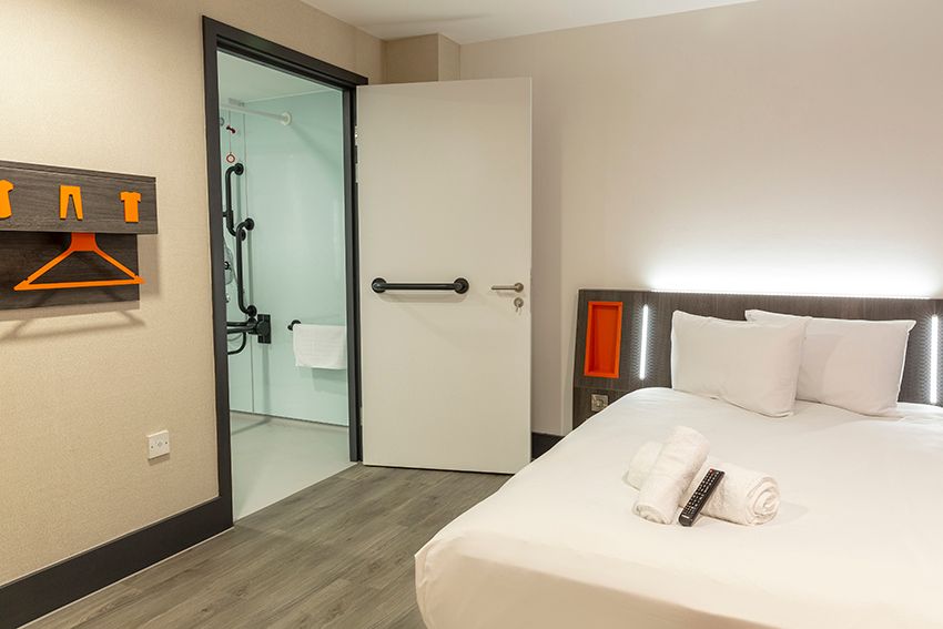 Accessible easyHotel Reading room featuring a crisp white bed, modern headboard with orange accents, and an open door revealing an accessible bathroom with grab bars. A wooden coat rack is on the left wall.