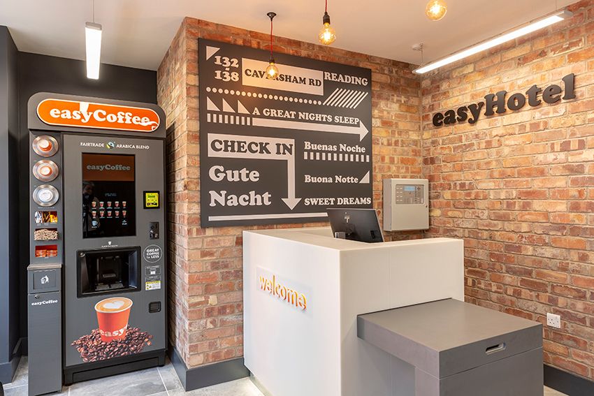 easyHotel Reading hotel lobby with a modern reception desk, an easyCoffee vending machine, and a large black sign on a brick wall displaying 'CHECK IN' and 'READING' with directions to 132-138 Caversham Road.