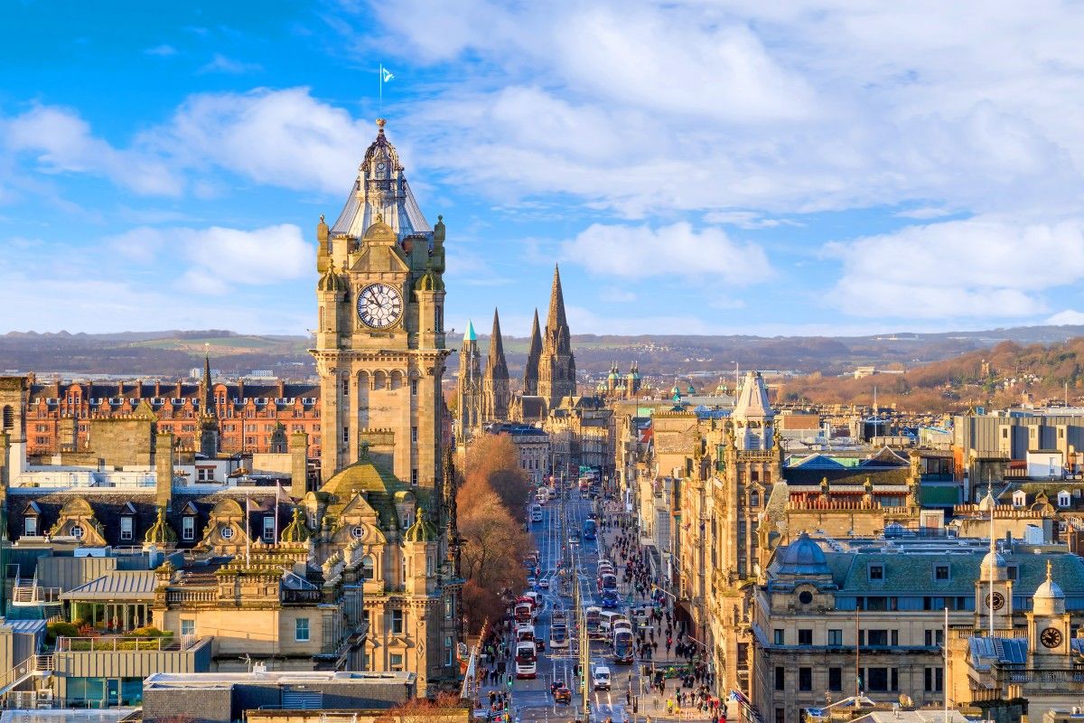 Panoramic view of Edinburgh's bustling Princes Street, featuring an iconic clock tower on the left, leading towards historic spires and buildings under a bright blue sky.
