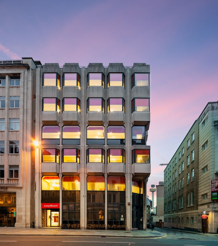 EasyHotel Liverpool building exterior at dusk, showcasing its Brutalist concrete facade and illuminated windows under a purple and pink sky, with the Radio City Tower visible.