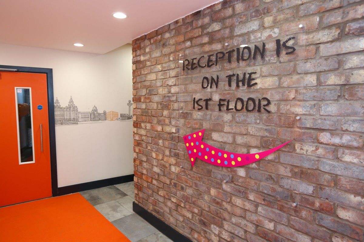 Modern hotel entrance hallway with an exposed brick wall displaying "RECEPTION IS ON THE 1ST FLOOR" text and a bright pink arrow. A mural of the Liverpool city skyline is visible on the adjacent white wall, next to a vibrant orange door and floor.