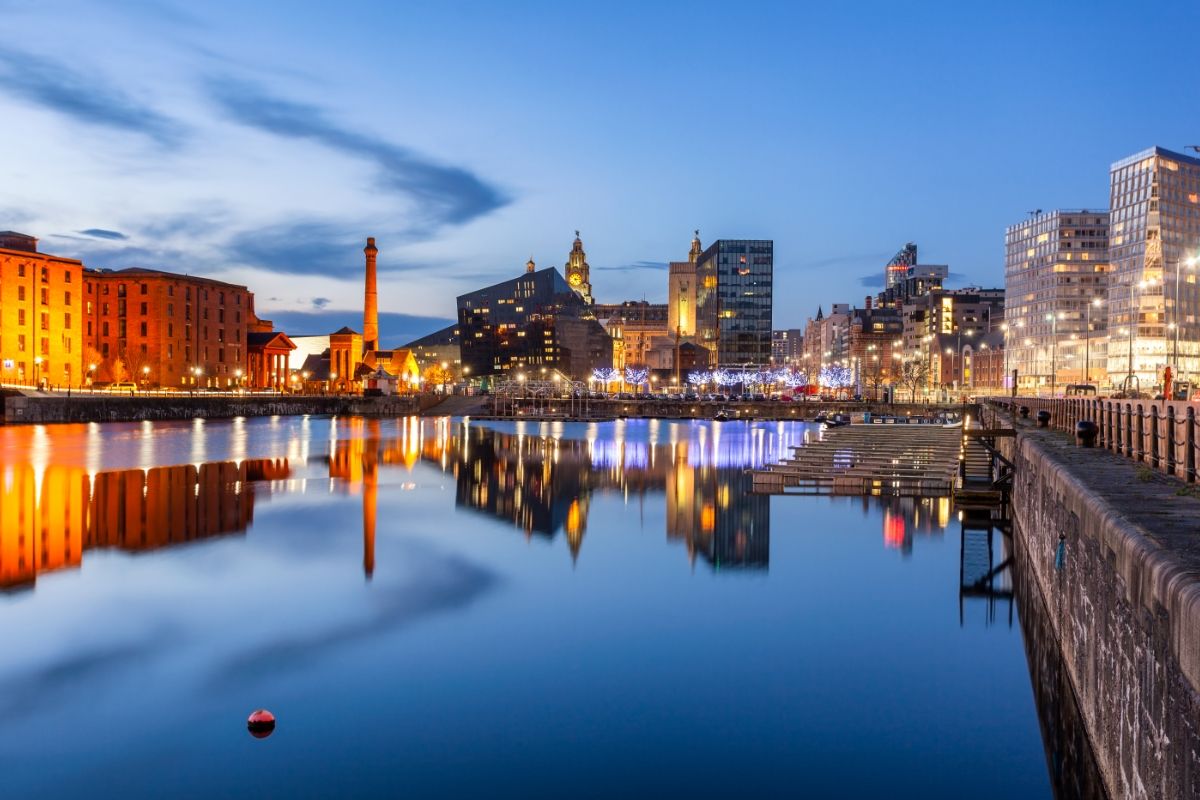 Stunning Liverpool waterfront cityscape at dusk, featuring illuminated historic Albert Dock buildings, modern architecture, and the iconic Liver Building reflecting in the calm water.