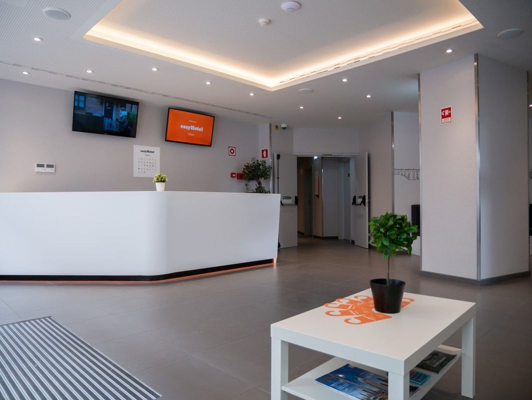 Bright, modern easyHotel reception lobby featuring a white curved desk, easyHotel branding on a screen and coffee table, light grey tiled floor, and a suspended ceiling with strip lighting.