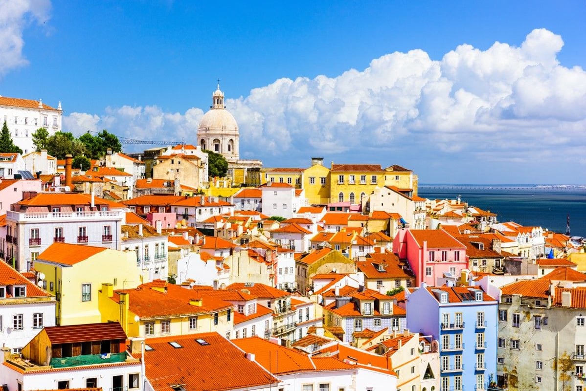Panoramic cityscape of Lisbon, Portugal, showcasing vibrant multi-colored historic buildings with terracotta roofs on hilly terrain. The white dome of the National Pantheon is visible, with the Tagus River and a distant bridge in the background under a br