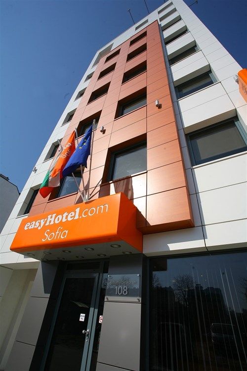 easyHotel Sofia exterior: modern building with orange and white facade, prominent easyHotel.com signage, and Bulgarian/EU flags under a clear blue sky.