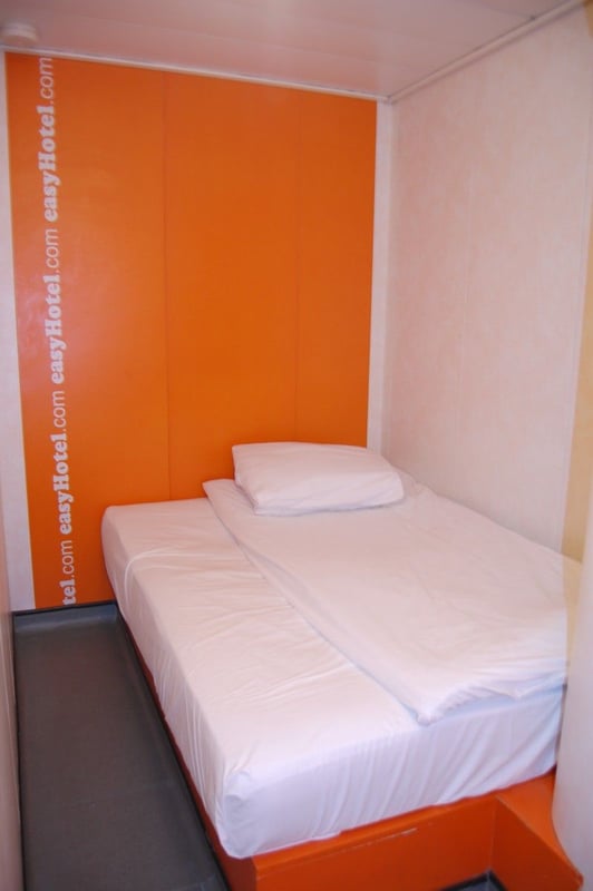Compact easyHotel standard double room featuring a white-linened bed and a vibrant orange wall prominently displaying the easyHotel.com brand logo.