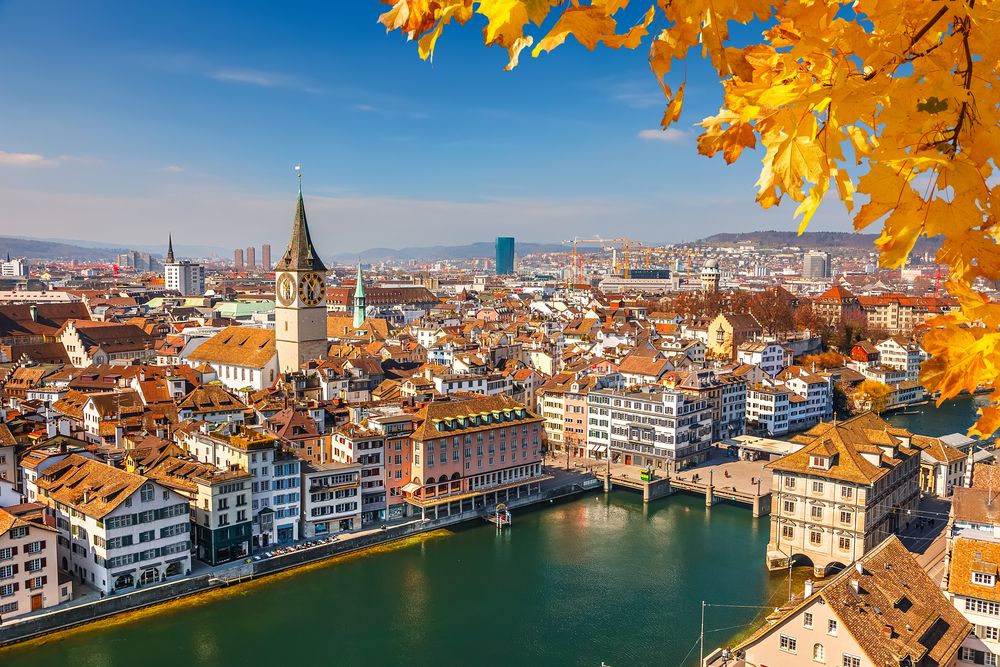 Autumnal panoramic view of Zurich, Switzerland, showcasing the Limmat River, historic Old Town buildings with terracotta roofs, and the iconic St. Peter Church clock tower, framed by vibrant golden yellow leaves under a clear blue sky.