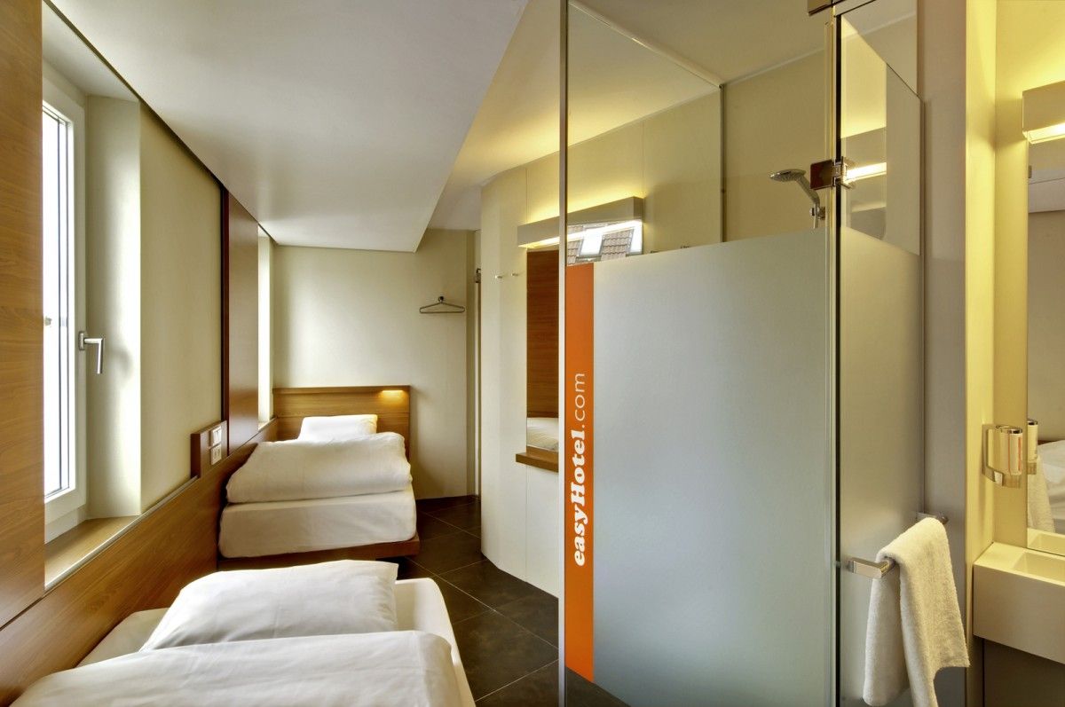 Compact easyHotel room in Berlin featuring two minimalist beds, a frosted glass en-suite shower, and modern design.